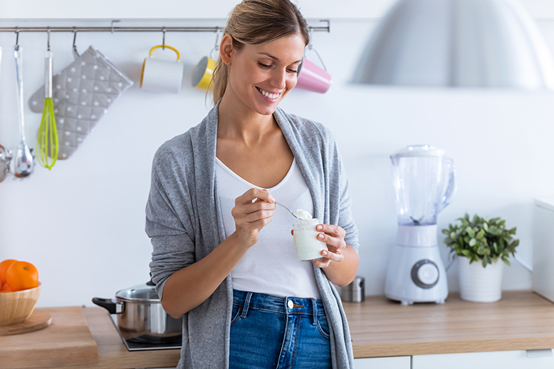 woman in her kitchen eating yogurt, one of 7 foods you can eat to burn belly fat