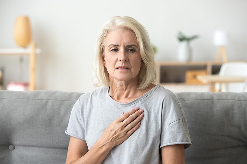 Older woman sitting on her couch in the living room feeling chest pain as a sign of heart attack.