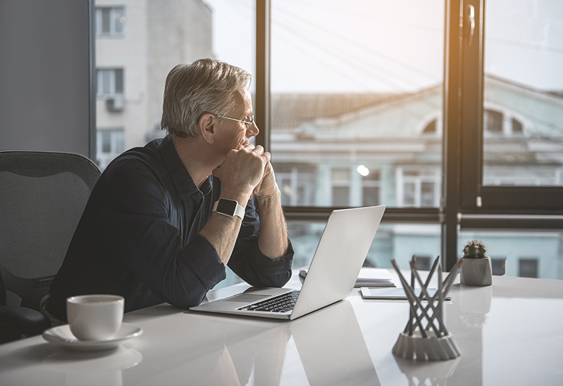 Businessman sitting at desk with laptop and looking out the window distracted at work.