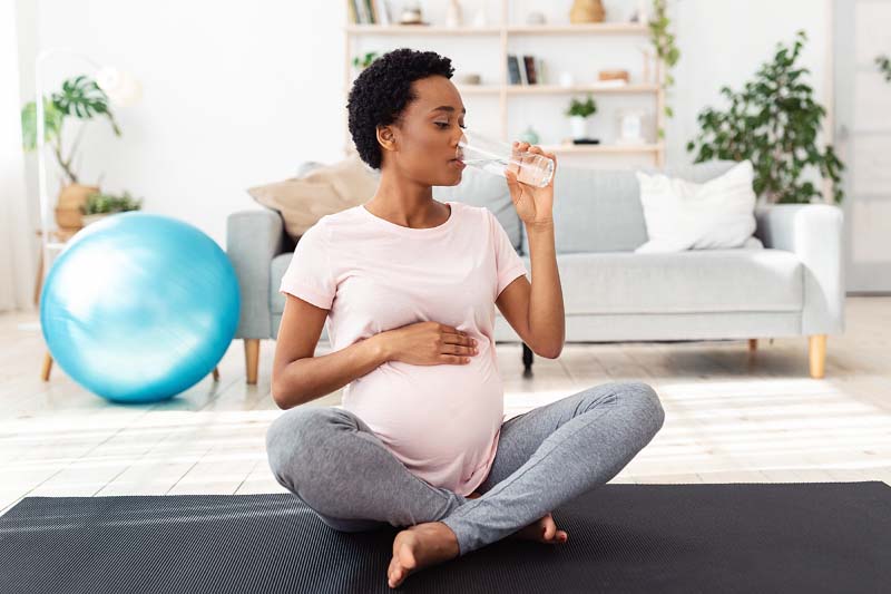 Pregnant woman drinking glass of water while exercising on yoga mat.