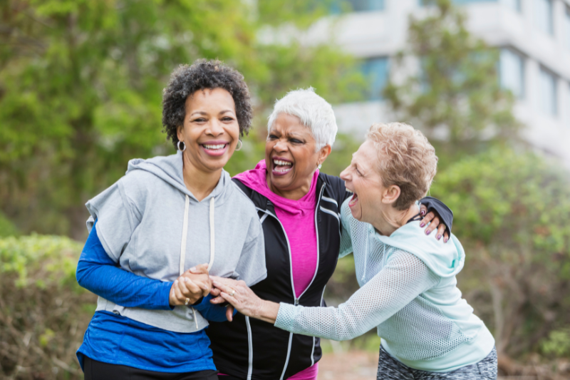 Three older women standing outside and wearing exercise clothes. They are holding each other and laughing.