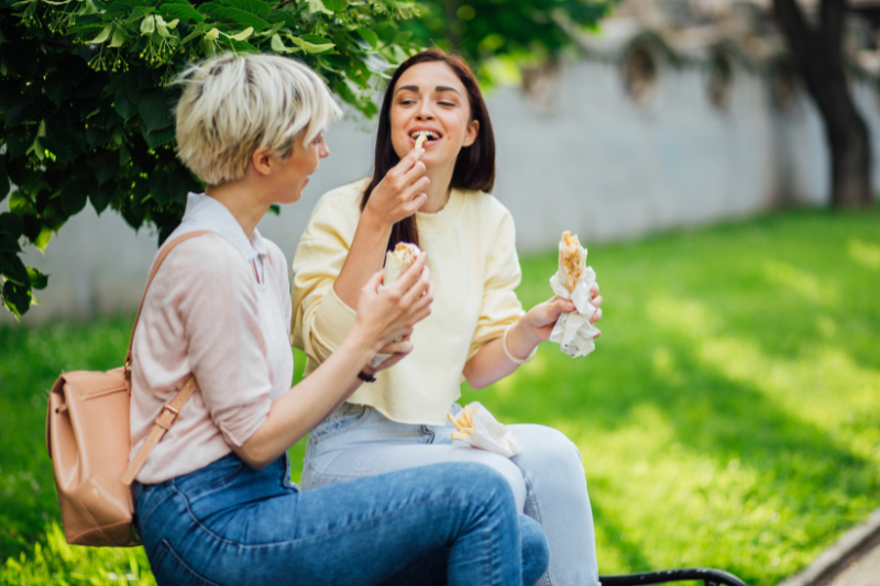  Two young women sitting on a bench, eating sandwiches and laughing.