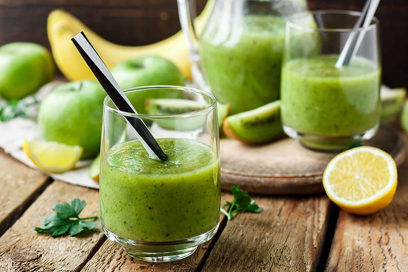 A glass of green goddess smoothie sits next to fruits on a table.